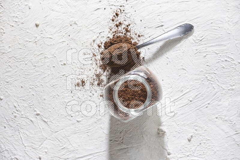A glass spice jar and a metal spoon full of cloves powder on textured white flooring