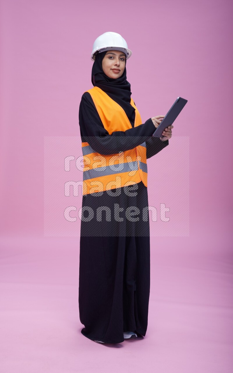 Saudi woman wearing Abaya with engineer vest and helmet standing working on tablet on pink background