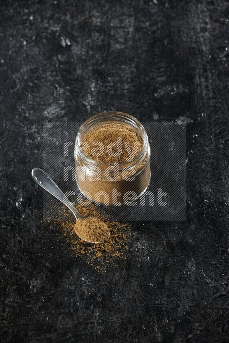 A glass jar and a metal spoon full of cumin powder on a textured black flooring