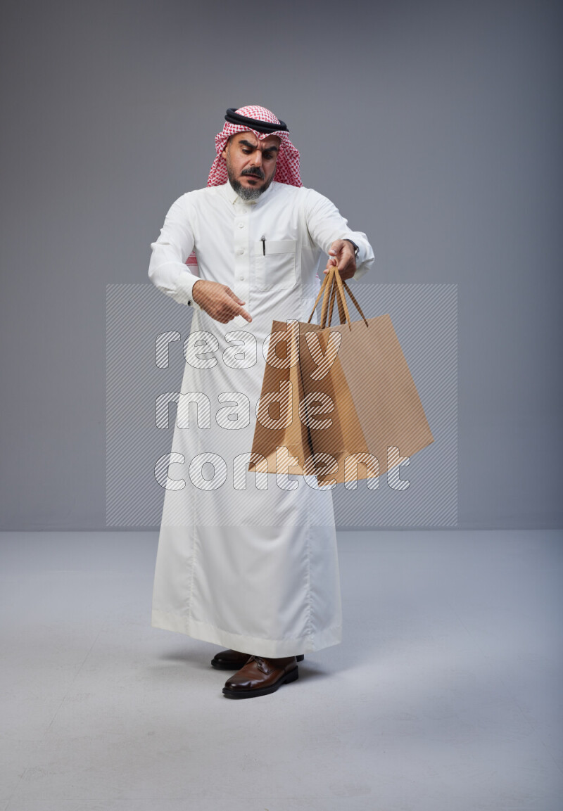 Saudi man Wearing Thob and red Shomag standing holding shopping bag on Gray background