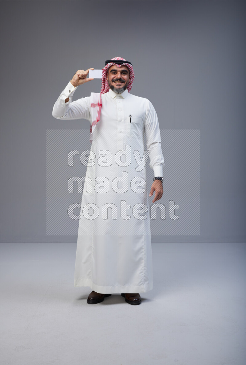Saudi man Wearing Thob and red Shomag standing holding ATM card on Gray background