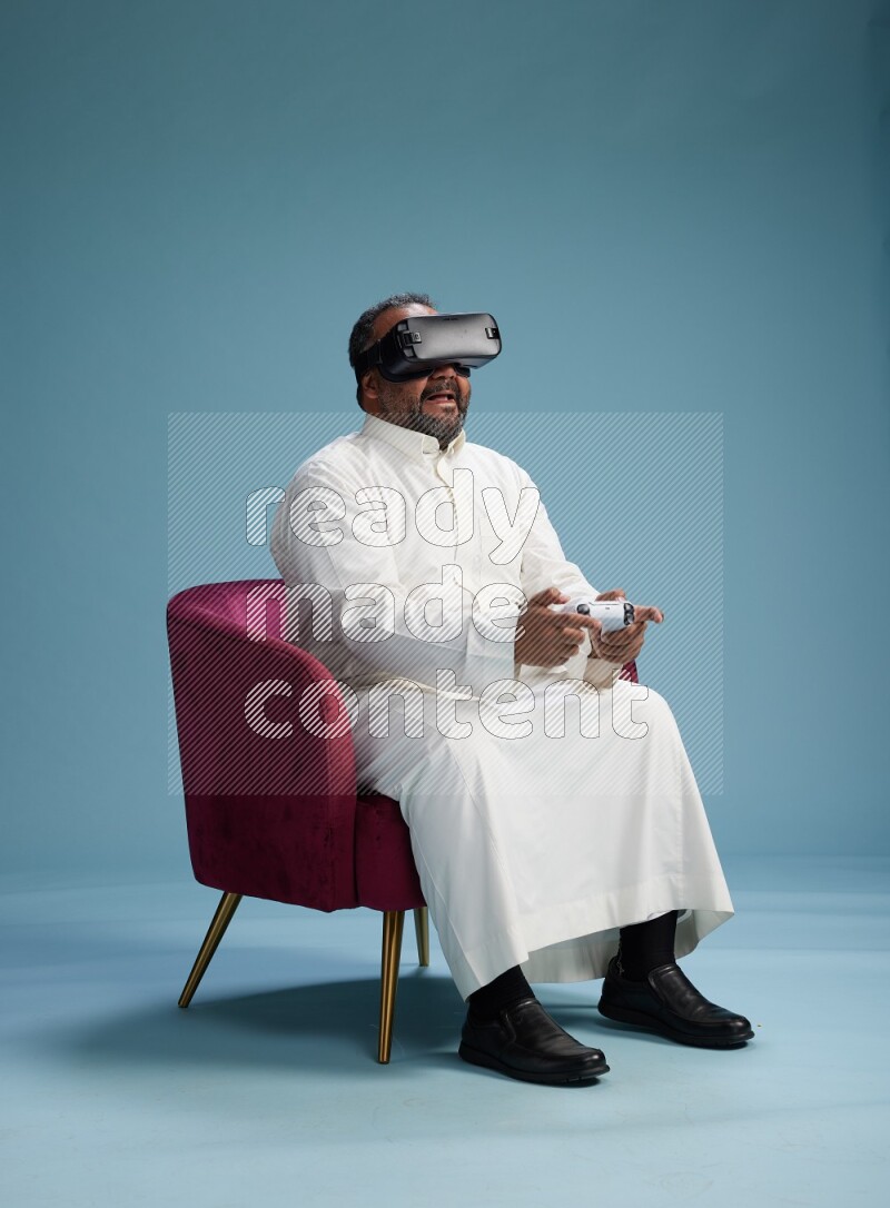 Saudi Man without shimag sitting on chair  gaming with VR and joystick on blue background