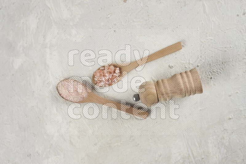 2 wooden spoons filled with fine and coarse salt with wooden grinder beside them on white background