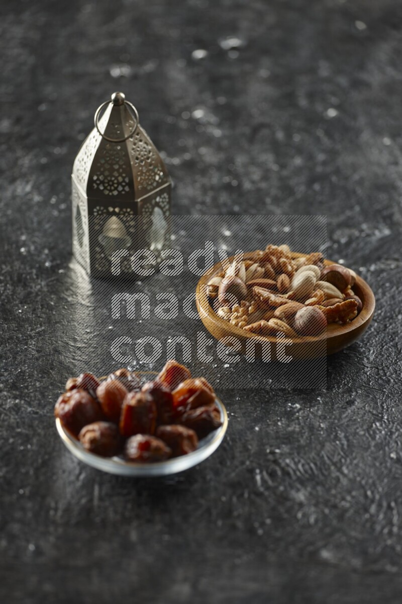 A silver lantern with different drinks, dates, nuts, prayer beads and quran on textured black background