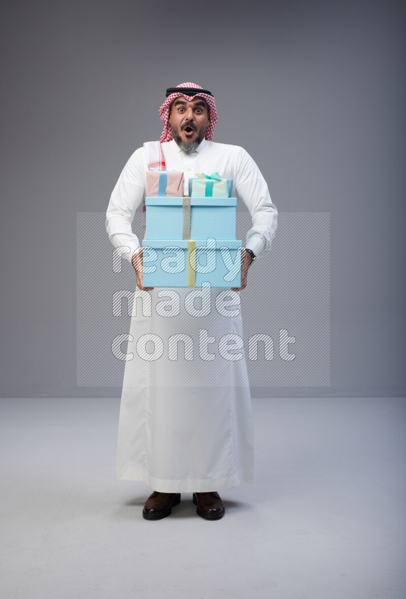 Saudi man Wearing Thob and red Shomag standing holding gift box on Gray background