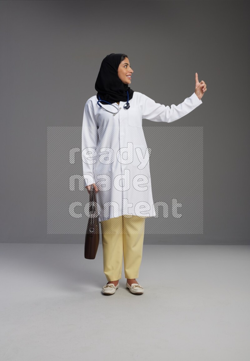 Saudi woman wearing lab coat with stethoscope standing holding bag on Gray background