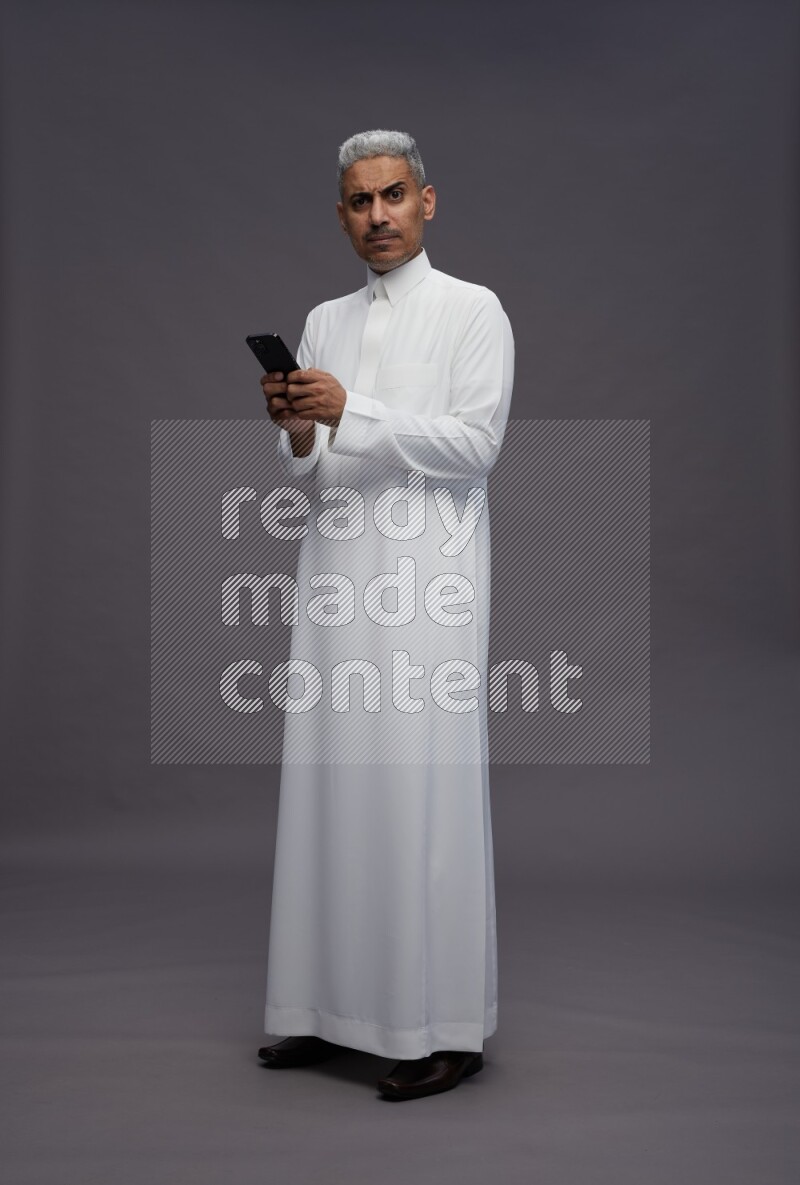 Saudi man wearing thob standing texting on phone on gray background