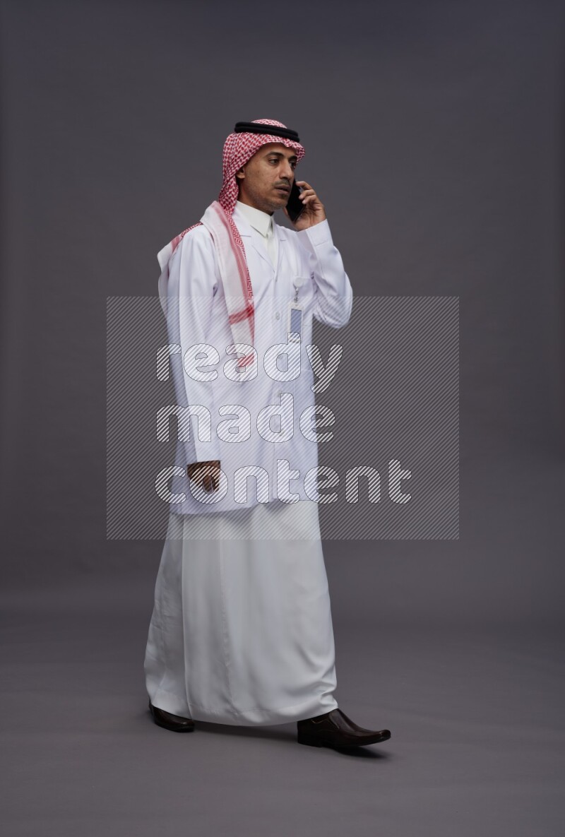 Saudi man wearing thob with lab coat and shomag with pocket employee badge standing talking on phone on gray background