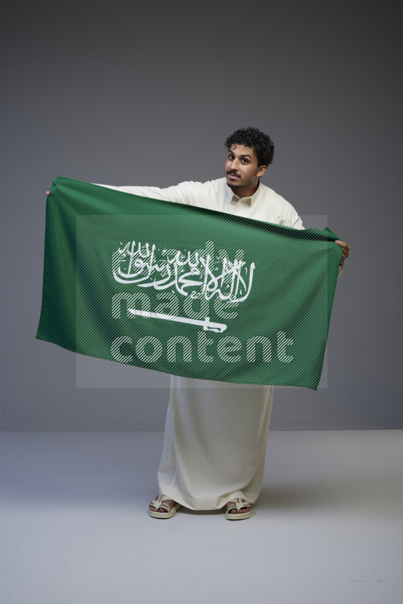 A Saudi man standing wearing thob holding big Saudi flag on gray background