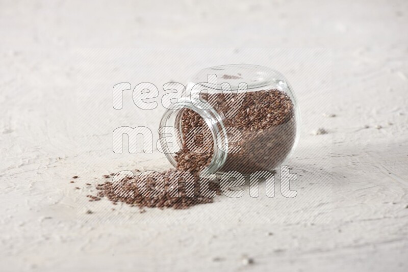 A glass spice jar full of flax seeds flipped and seeds spread out on a textured white flooring
