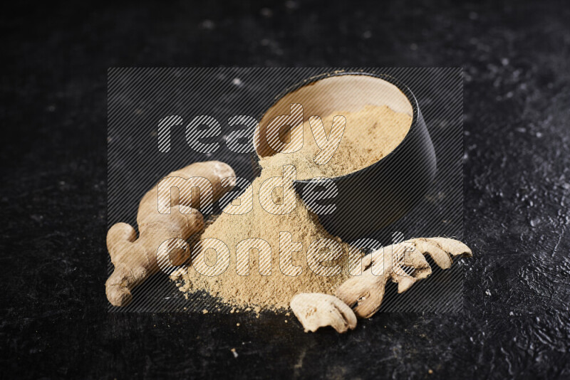 A black pottery bowl full of ground ginger powder with fallen powder from it on black background