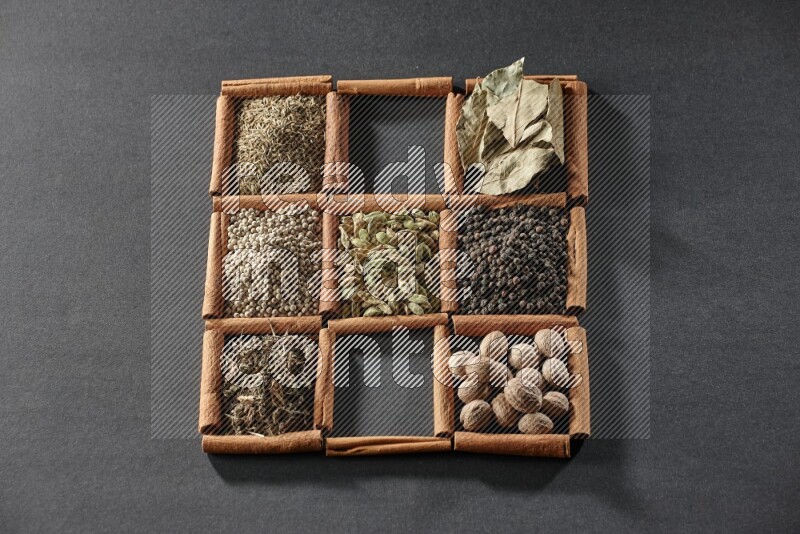 9 مربعات من أعواد القرفة ممتلئة بالهيل في المنتصف محاطة بجوزة الطيب والفلفل الأسود وورق الغار والبهارات والكمون والفلفل الأبيض والريحان المجفف والملح على أرضية سوداء