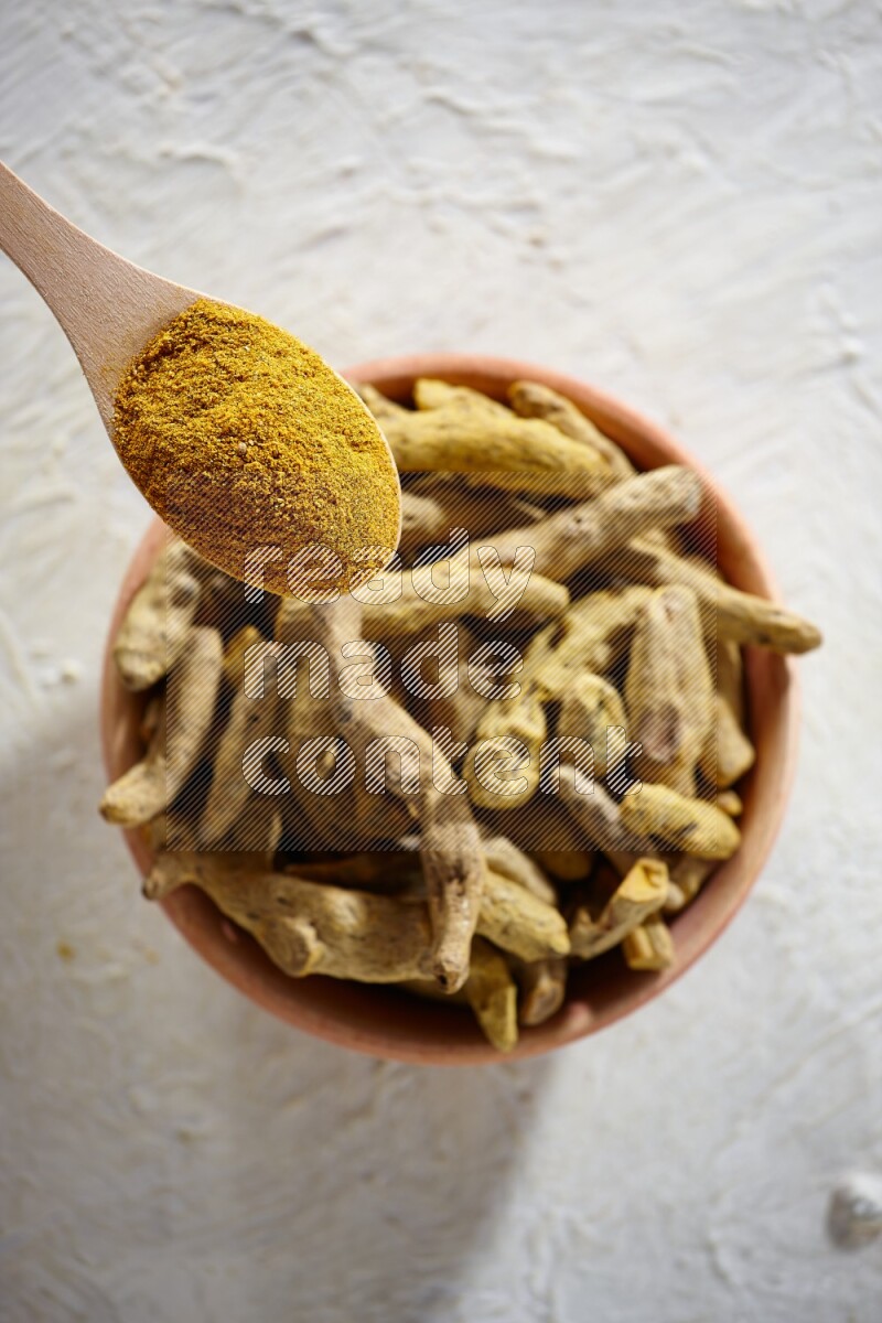 A wooden spoon full of turmeric powder above a wooden bowl full of dried turmeric whole fingers on a textured white flooring