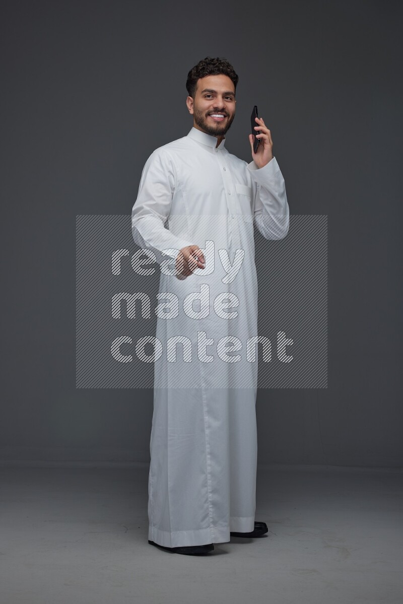 A Saudi man wearing Thobe and talking in the phone while standing and making different poses eye level on a gray background