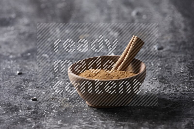 Wooden bowl full of cinnamon powder and a cinnamon stick on a textured black background