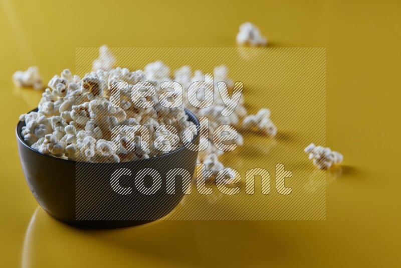 A black ceramic bowl full of popcorn with popcorn beside it on a yellow background in different angles