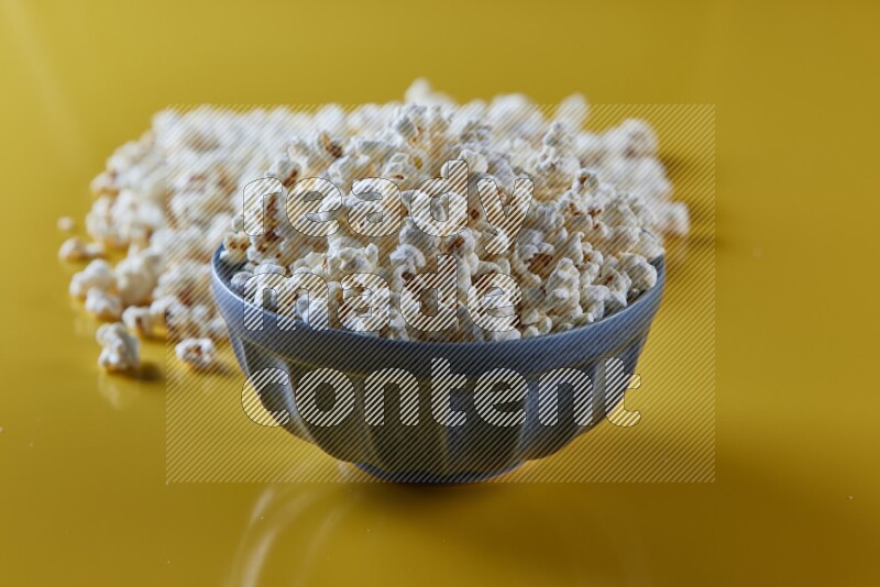 A blue pottery bowl full of popcorn with popcorn beside it on a yellow background in different angles