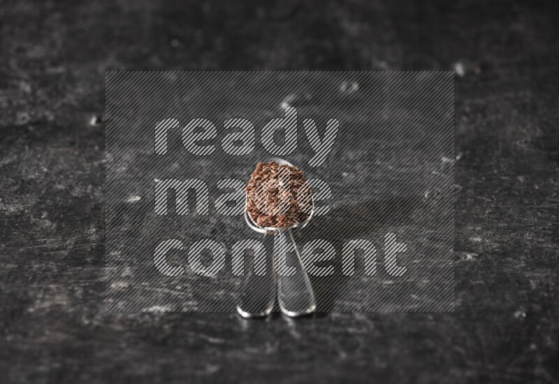 2 metal spoons full of flaxseeds on a textured black flooring