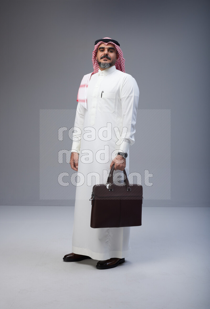 Saudi man Wearing Thob and red Shomag standing holding bag on Gray background