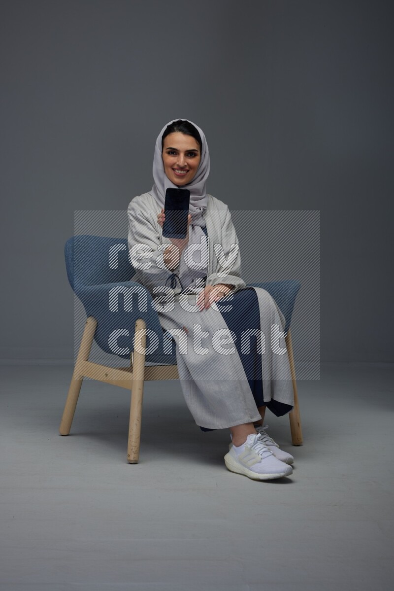 A Saudi woman wearing a light gray Abaya and head scarf sitting on a dark grey chair and showing the phone's screen eye level on a grey background