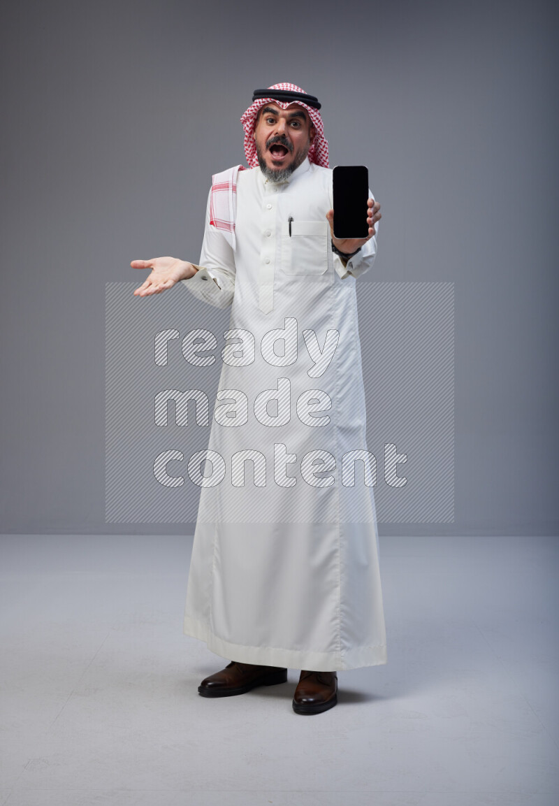 Saudi man Wearing Thob and red Shomag standing showing phone to camera on Gray background