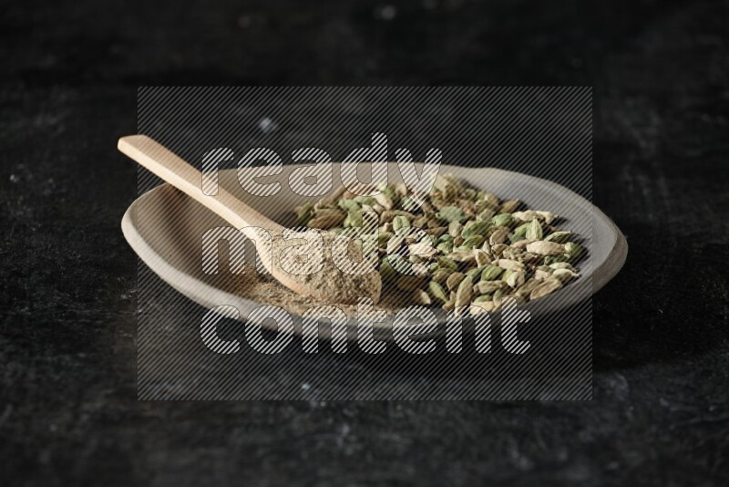 A plate filled with cardamom seeds and a wooden spoon full of cardamom powder on a textured black flooring