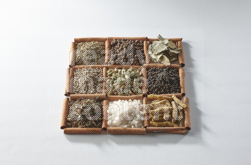 9 مربعات من أعواد القرفة ممتلئة بالهيل في المنتصف محاطة بأصابع الكركم والملح والريحان المجفف والفلفل الأبيض والكمون وبهار الفلفل الحلو وأوراق الغار والفلفل الأسود على أرضية بيضاء