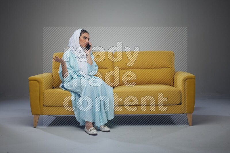 A Saudi woman wearing a light blue Abaya and a white head scarf sitting on a yellow sofa and talking in the phone while making different poses eye level on a grey background