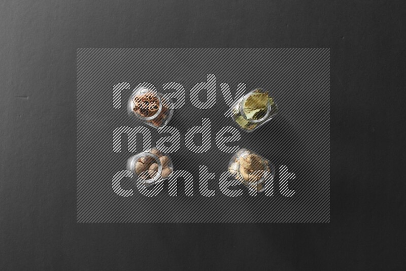 4 glass spice jars full of laurel bay leaves, ginger, cinnamon and nutmeg on black flooring