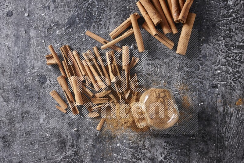 Herbal glass jar full cinnamon powder flipped and a metal spoon full of powder surrounded by cinnamon sticks on textured black background in different angles