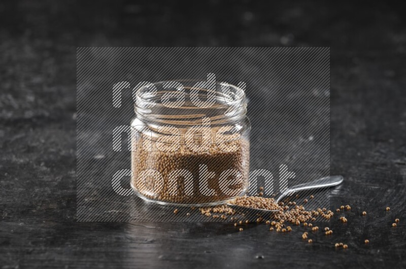 A glass jar and a metal spoon full of mustard seeds on a textured black flooring