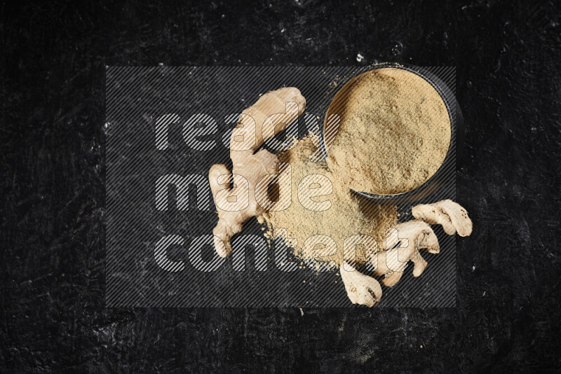 A black pottery bowl full of ground ginger powder with fallen powder from it on black background