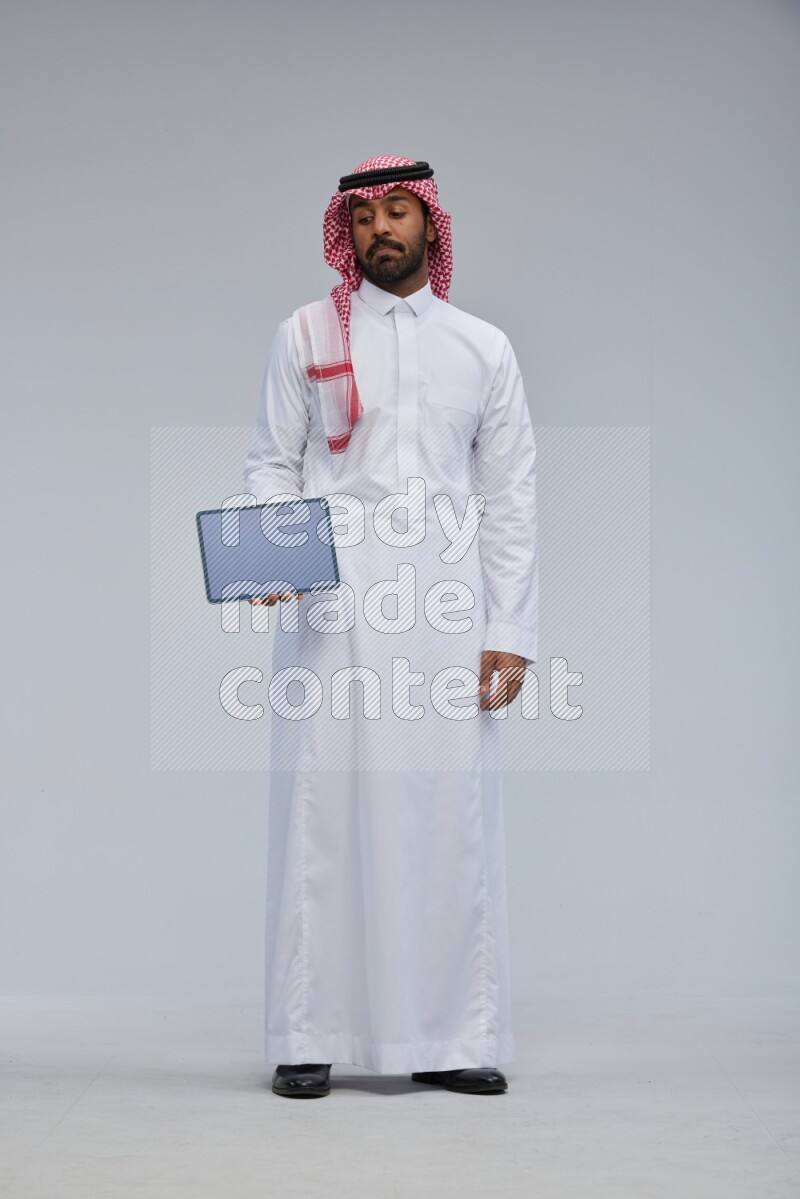 Saudi man Wearing Thob and shomag standing showing tablet to camera on Gray background