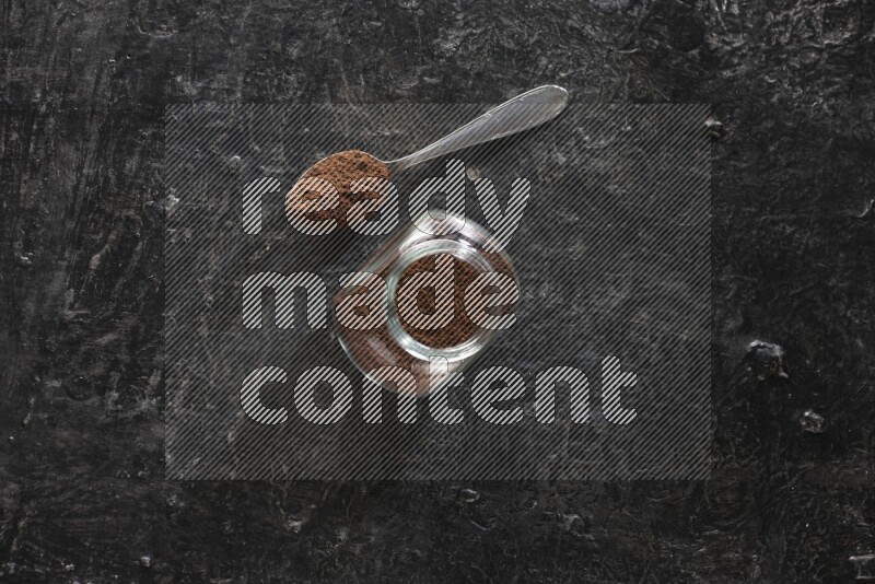 A glass spice jar and a metal spoon full of cloves powder on textured black flooring