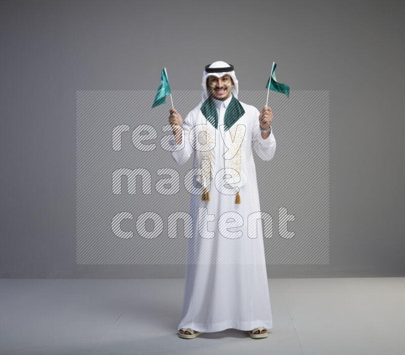 A saudi man standing wearing thob and white shomag with face painting and saudi flag scarf and holding small saudi flag on gray background