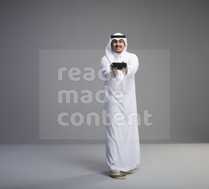 A Saudi man standing wearing thob and white shomag showing phone to camera on gray background