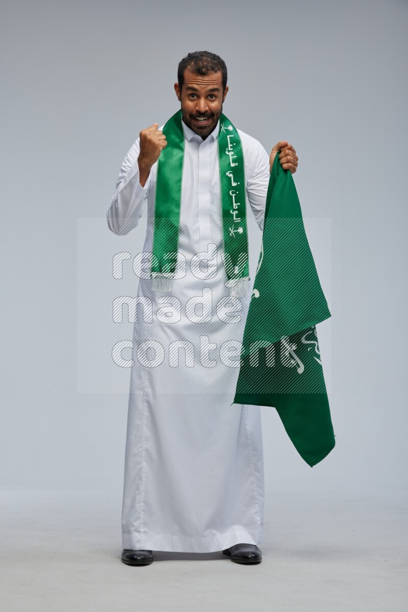 Saudi man wearing Thob standing holding Saudi flag on Gray background