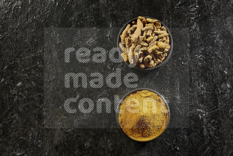 2 pottery black bowls, one full of turmeric powder and the other full of dried turmeric whole fingers on a textured black flooring