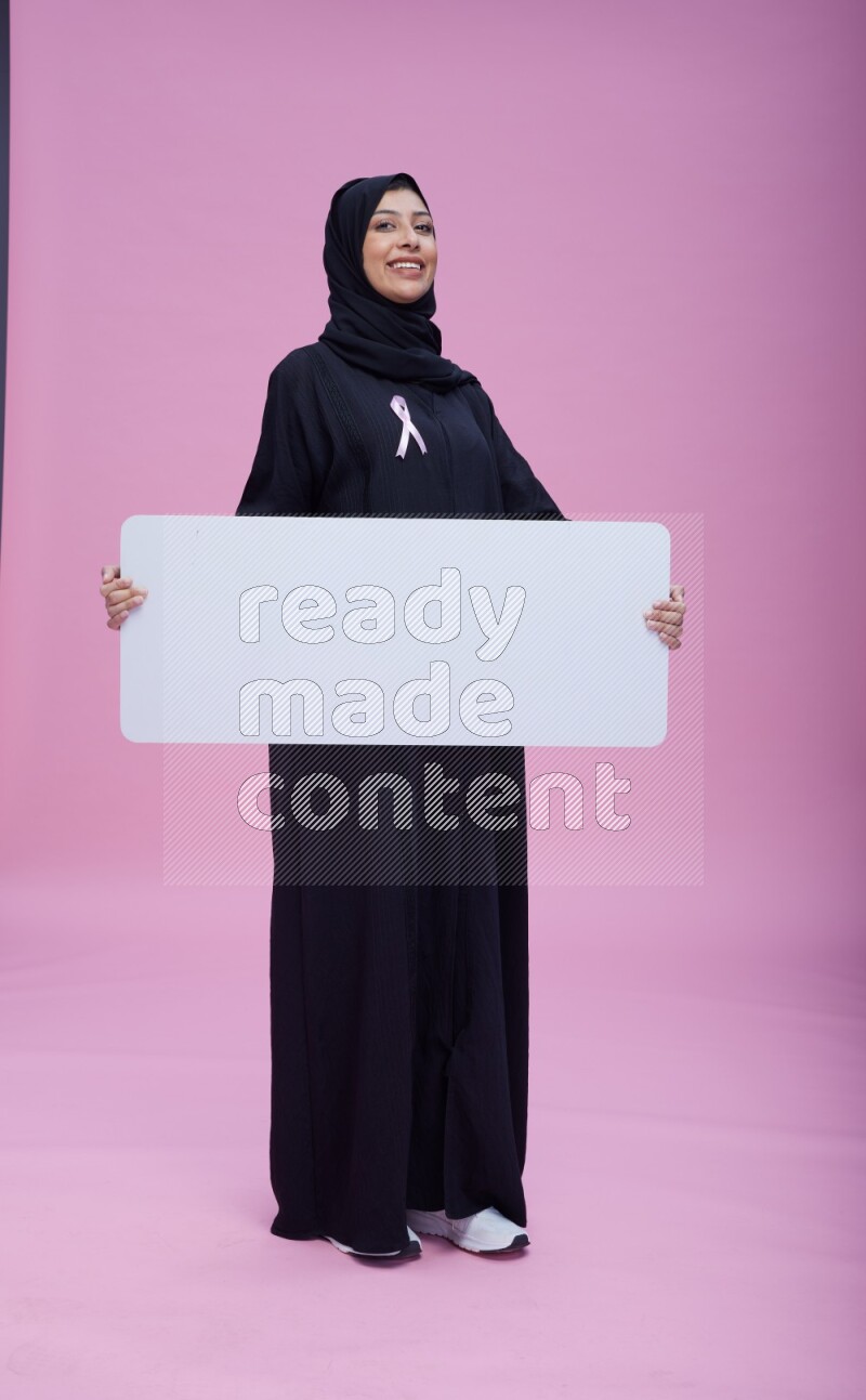 Saudi woman wearing pink ribbon on Abaya standing holding board on pink background