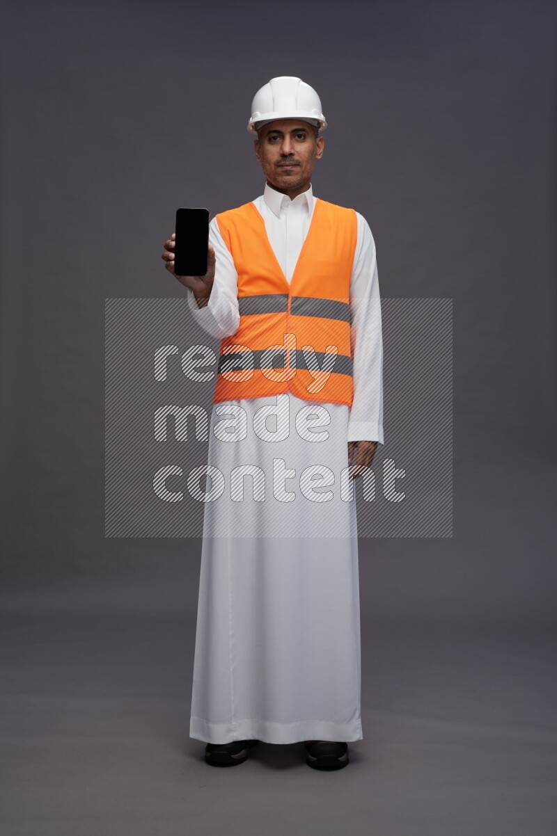 Saudi man wearing thob with engineer vest standing showing phone to camera on gray background