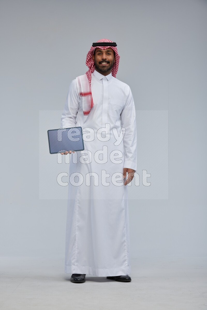 Saudi man Wearing Thob and shomag standing showing tablet to camera on Gray background