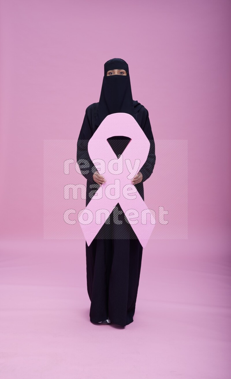 Saudi woman wearing abaya and niqab standing holding awareness ribbon on pink background