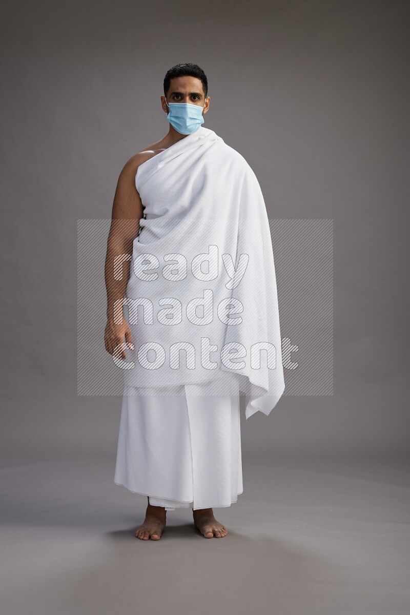A man wearing Ehram with face mask Standing Interacting with the camera on gray background