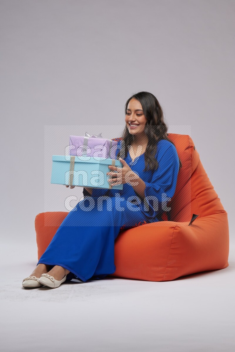 A woman sitting on an orange beanbag wearing Jalabeya holding a gift box
