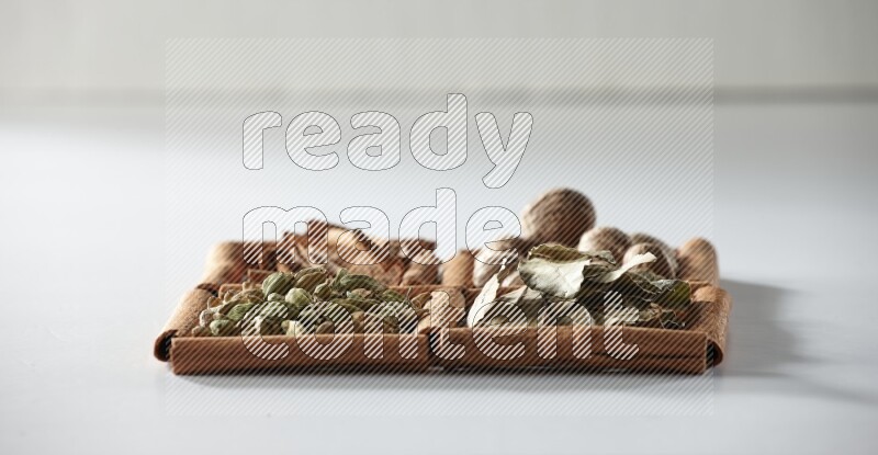 4 مربعات من أعواد القرفة ممتلئة بجوزة الطيب وأوراق الغار وحبوب الهيل والقرفة على أرضية بيضاء