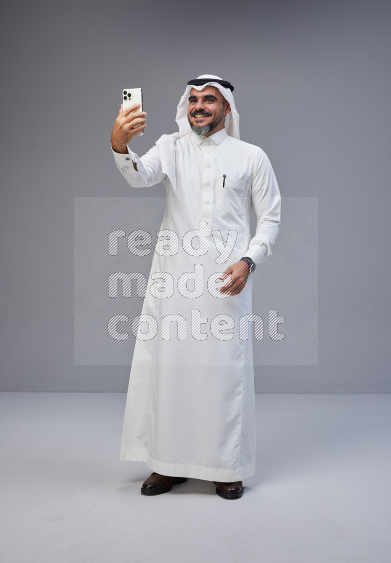 Saudi man Wearing Thob and white Shomag standing taking selfie on Gray background