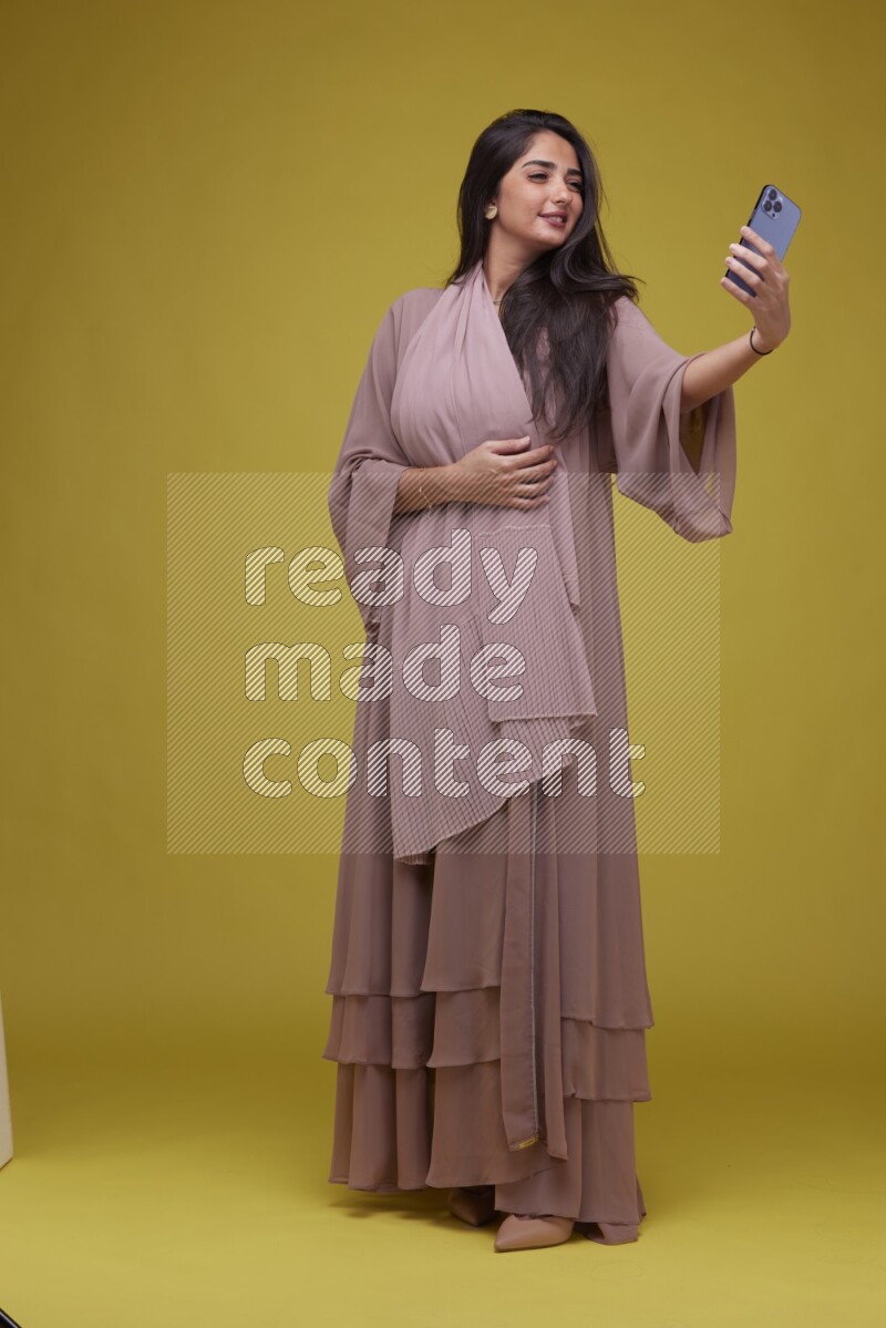 A woman Taking a Selfie on a Yellow Background wearing Brown Abaya