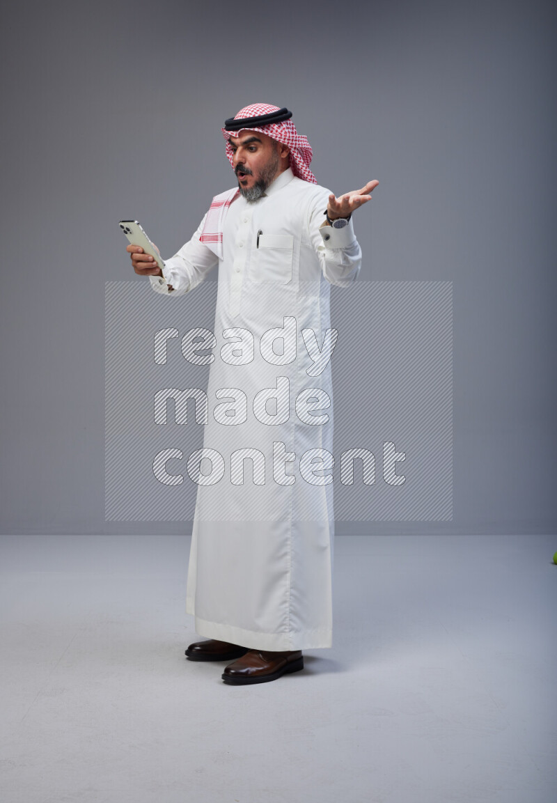 Saudi man Wearing Thob and red Shomag standing texting on phone on Gray background