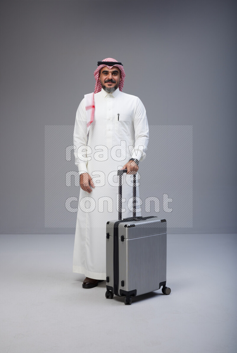 Saudi man wearing Thob and red Shomag standing holding Travel bag on Gray background
