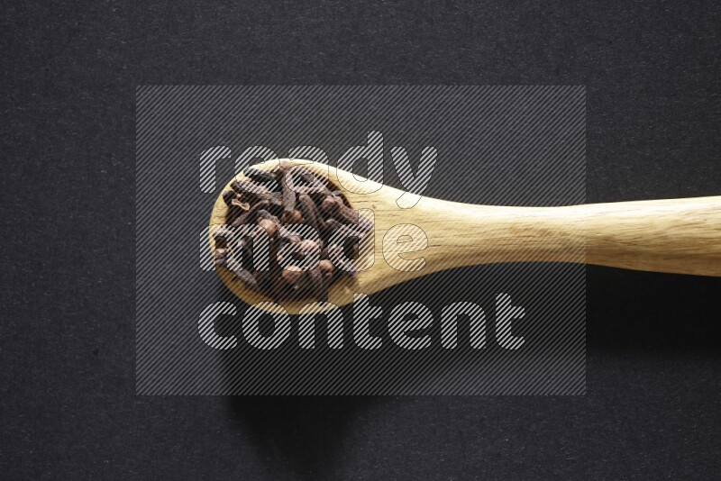 A wooden spoon full of cloves on a black flooring